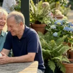 A senior man and woman in their 70s laughing together over coffee at an outdoor garden cafe during a sunny afternoon.