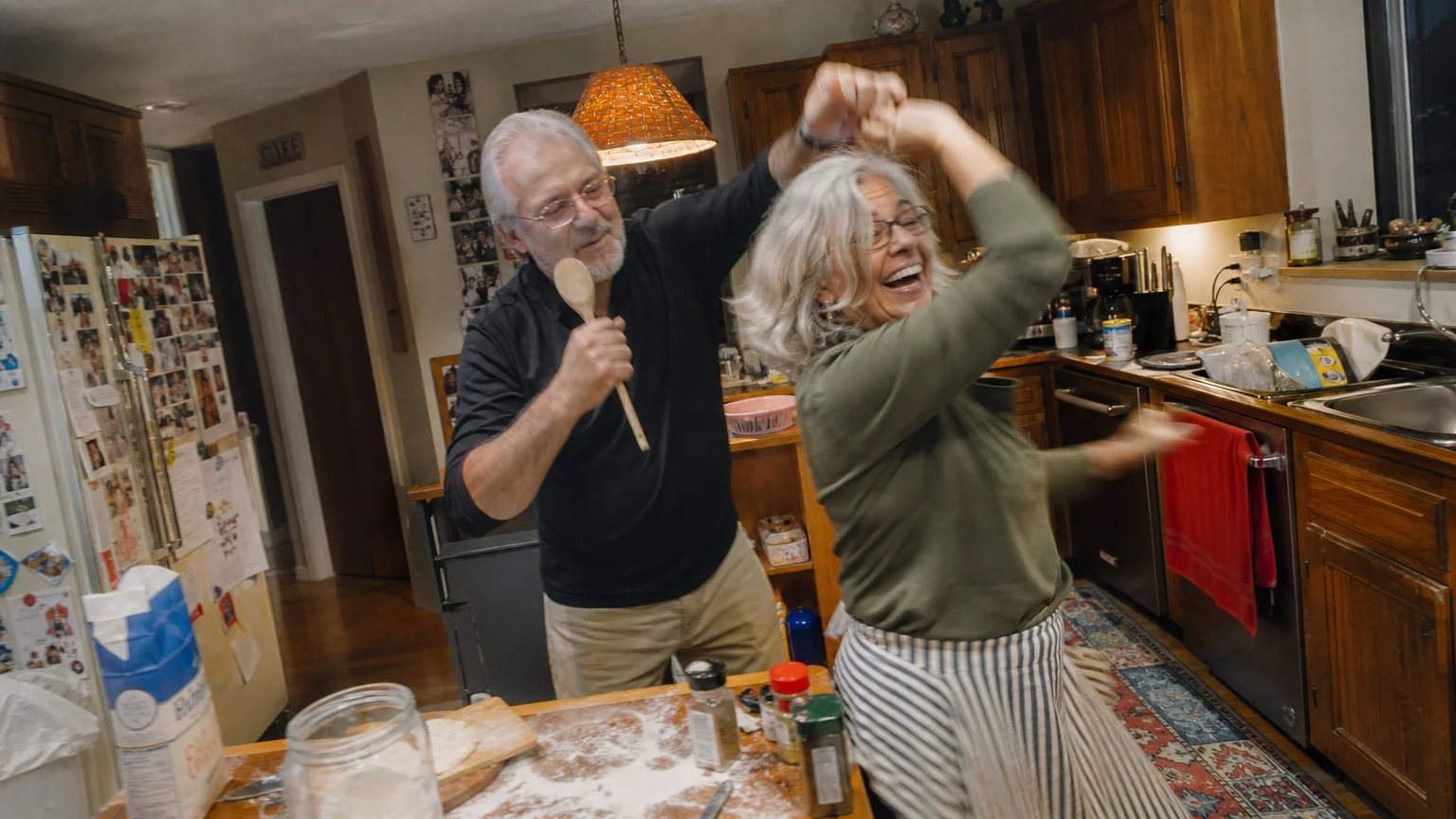 A candid, slightly blurred photo of a senior couple dancing and laughing in their messy kitchen during dinner prep.