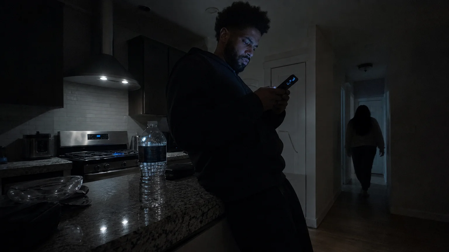 A person leans against a dark kitchen counter at night, frustration on their face as they type a message on a glowing phone.
